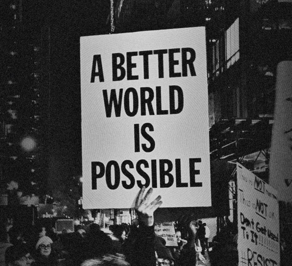 Protesters at night; one holds a large hand-lettered sign reading "A better world is possible."